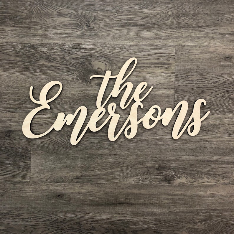 May include: The Emersons family name in cursive script, cut out of wood and painted white, against a dark wood background.