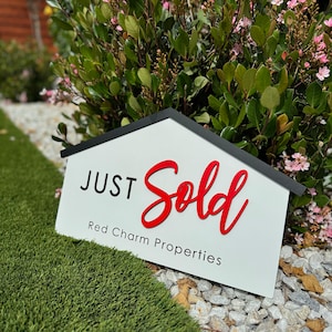 May include: A white sign with black trim shaped like a house reads "JUST Sold" in red cursive lettering. Below the text is "Red Charm Properties" in black lettering.