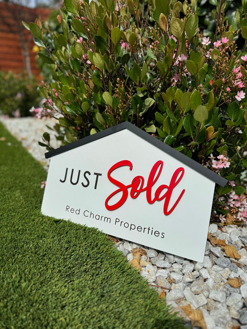 May include: A white sign with black trim shaped like a house reads "JUST Sold" in red cursive lettering. Below the text is "Red Charm Properties" in black lettering.