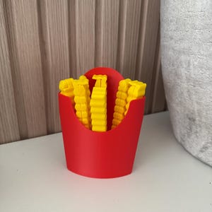 May include: A red container filled with yellow, french fry-shaped objects. The container is a solid red color and has a curved top. The yellow objects are arranged to look like french fries.