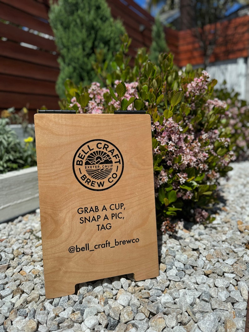 May include: A wooden sign with a black logo that says "Bell Craft Brew Co. Exeter, Calif." and the text "Grab a cup, snap a pic, tag @bell_craft_brewco".