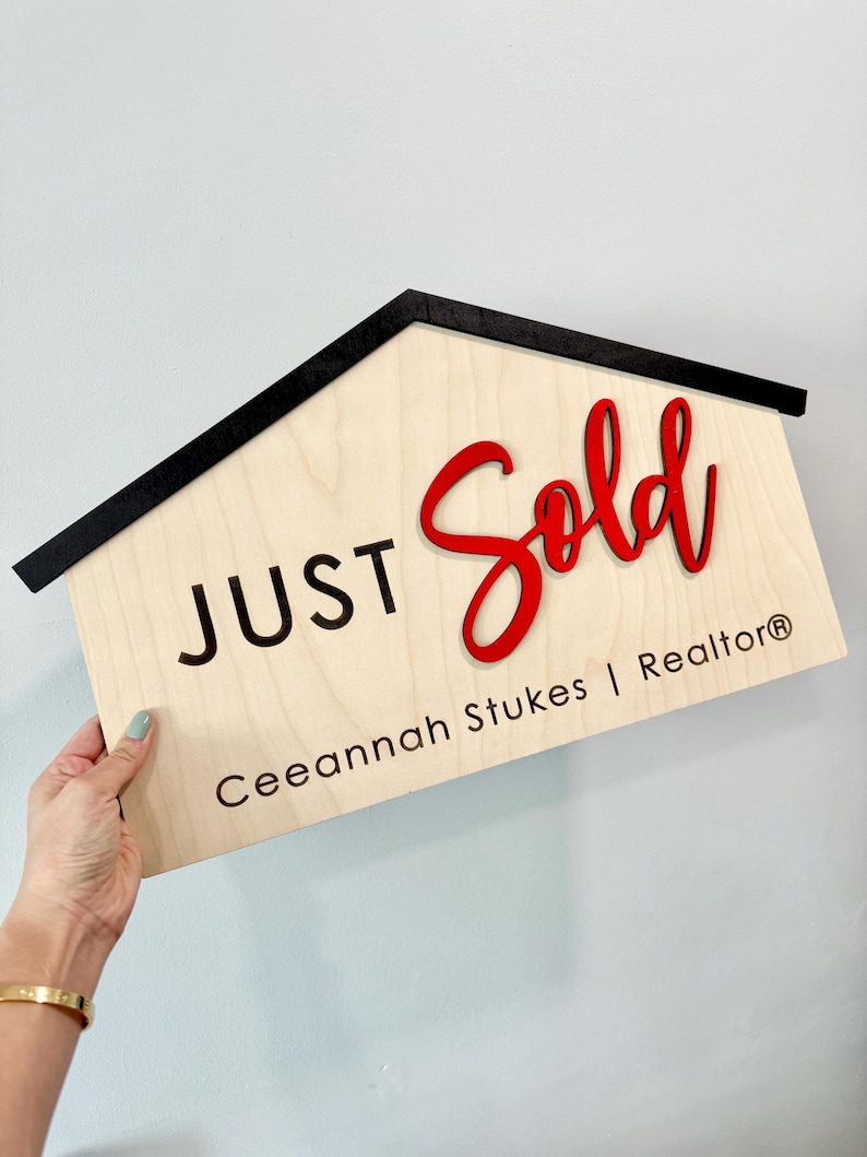 May include: A wooden house-shaped sign with a black border and the words "JUST SOLD" in red and black lettering. The name "Ceeannah Stukes | Realtor&reg;" is written below in black lettering.