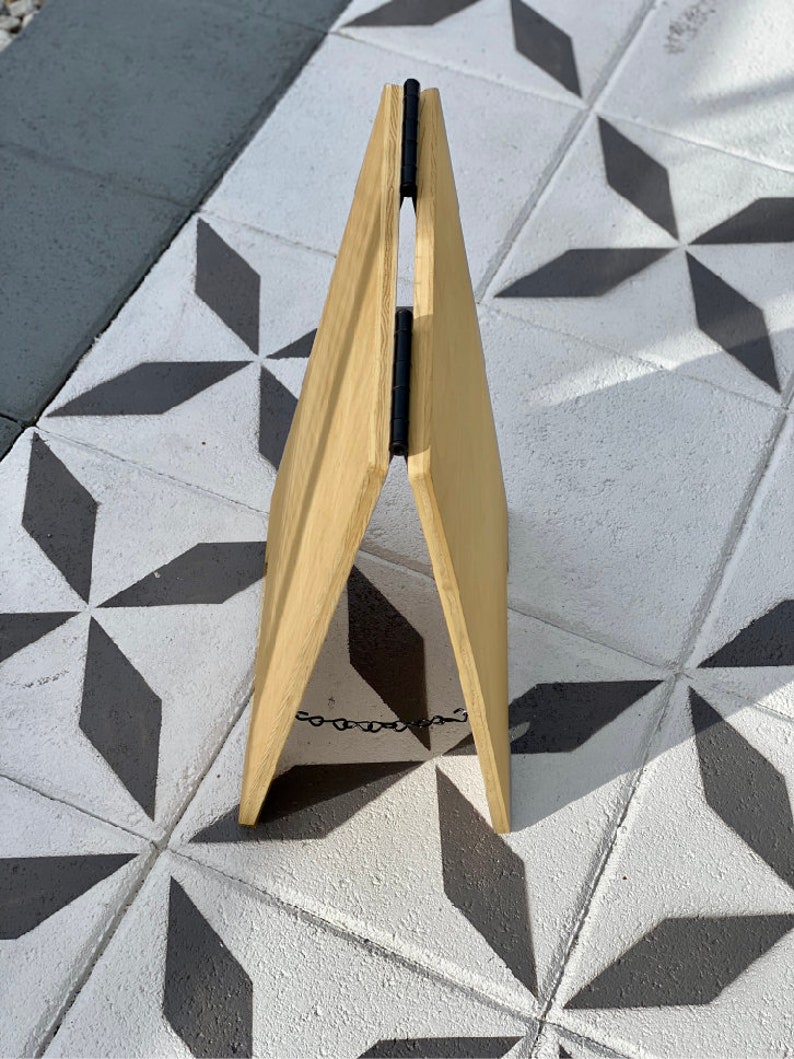 May include: A wooden A-frame sign with a black chain attached to the bottom. The sign is standing on a tiled floor with a geometric pattern.