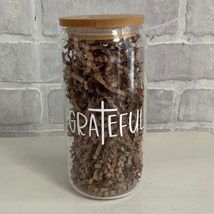 Puede incluir: Un tarro de vidrio transparente con tapa de madera lleno de papel de seda marrón. El tarro tiene la palabra "GRATEFUL" escrita en blanco con una cruz blanca en el medio de la palabra.