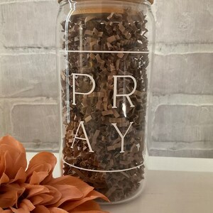 Puede incluir: Un tarro de vidrio transparente con tapa de bambú lleno de papel de seda marrón. El tarro tiene la palabra "PRAY" escrita en blanco.
