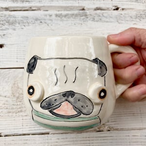 May include: A white ceramic mug with a hand-painted pug face. The pug has black eyes, a black nose, and a pink mouth. The mug has a green and white striped design around the bottom.