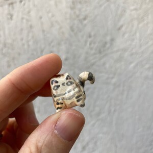 May include: A small, hand-carved wooden cat figurine. The cat is painted with black and brown stripes and spots on a cream-colored base. The tail is curled up. The figurine is held in a person's hand.