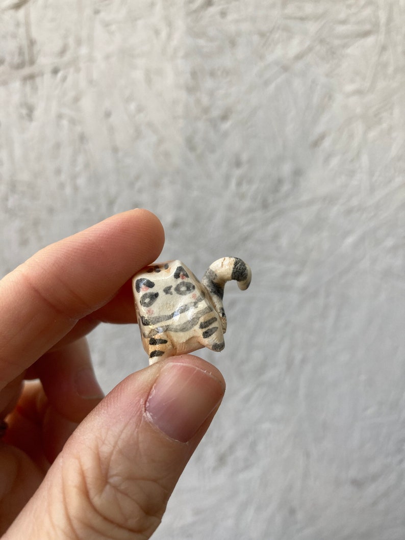 May include: A small, hand-carved wooden cat figurine. The cat is painted with black and brown stripes and spots on a cream-colored base. The tail is curled up. The figurine is held in a person's hand.