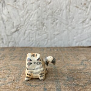 May include: A small, handmade ceramic cat figurine. The cat is painted with a cream base, black stripes, and pink cheeks. The tail is curled and striped. The figurine is set on a wooden surface.