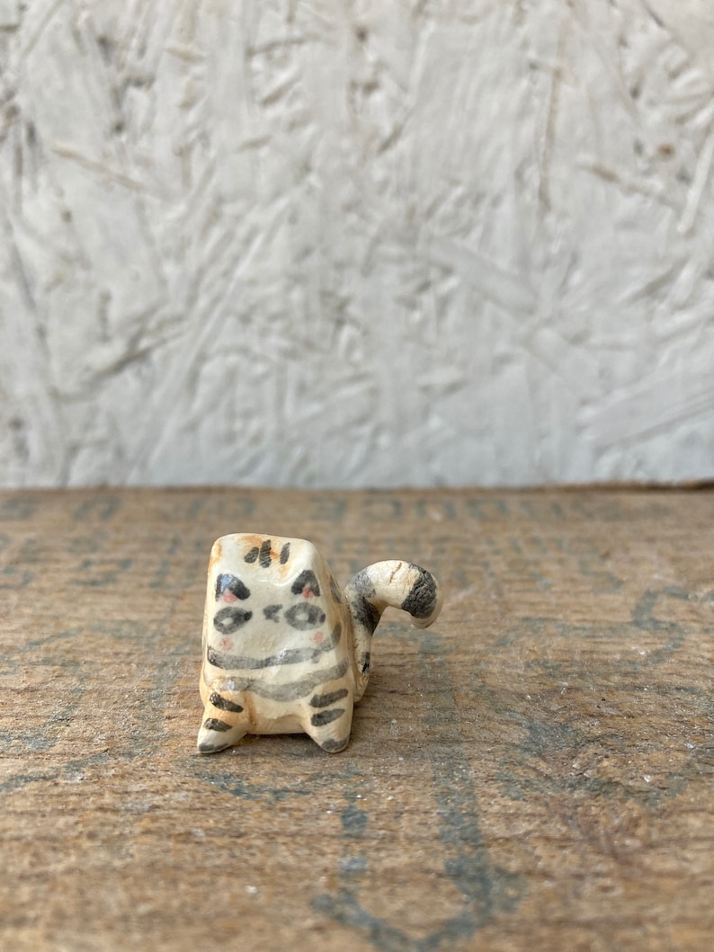 May include: A small, handmade ceramic cat figurine. The cat is painted with a cream base, black stripes, and pink cheeks. The tail is curled and striped. The figurine is set on a wooden surface.