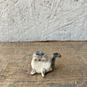 May include: A small, handmade ceramic cat figurine. The cat is primarily white with gray and black markings, including a gray head and tail. It has small pink accents on its cheeks and closed eyes, giving it a relaxed appearance. The figurine is on a wooden surface.