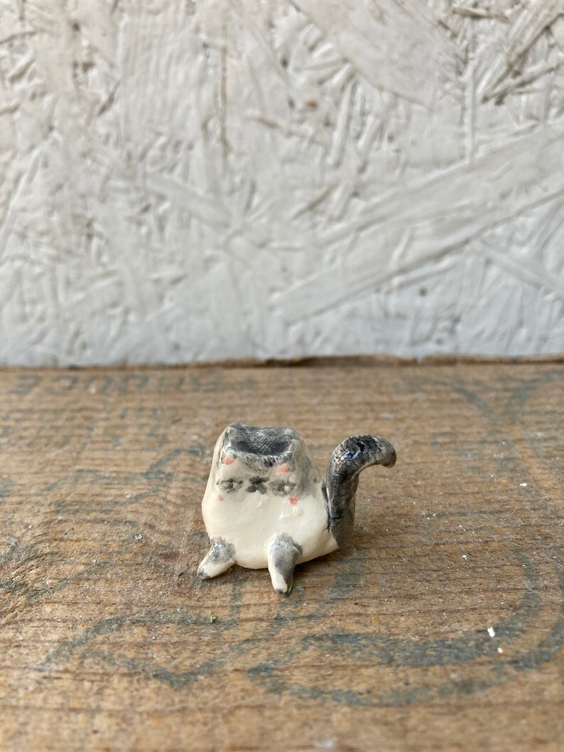 May include: A small, handmade ceramic cat figurine. The cat is primarily white with gray and black markings, including a gray head and tail. It has small pink accents on its cheeks and closed eyes, giving it a relaxed appearance. The figurine is on a wooden surface.