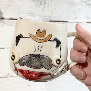 May include: White ceramic mug with a hand-painted pug wearing a cowboy hat and bandana. The mug has the words "How" written on the side.