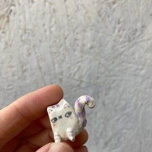 May include: A small, handmade ceramic cat figurine. The cat is white with purple markings on its head, legs, and tail. It has black eyes and pink cheeks. The tail is curled. The figurine is held in a hand.