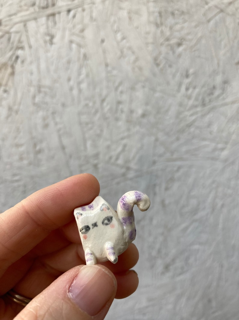 May include: A small, handmade ceramic cat figurine. The cat is white with purple markings on its head, legs, and tail. It has black eyes and pink cheeks. The tail is curled. The figurine is held in a hand.