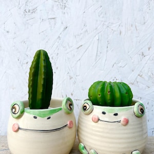 May include: Two ceramic frog-shaped planters, each holding a green cactus. The planters are off-white with green and pink accents, featuring frog-like eyes, cheeks, and feet. One planter has a tall, thin cactus, the other a rounder one.