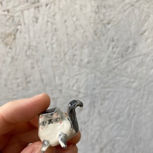 May include: A small, handmade ceramic planter in the shape of a turtle. The planter is primarily white with black and gray accents, including the turtle's head and tail. The planter has three small feet and a small opening for planting.