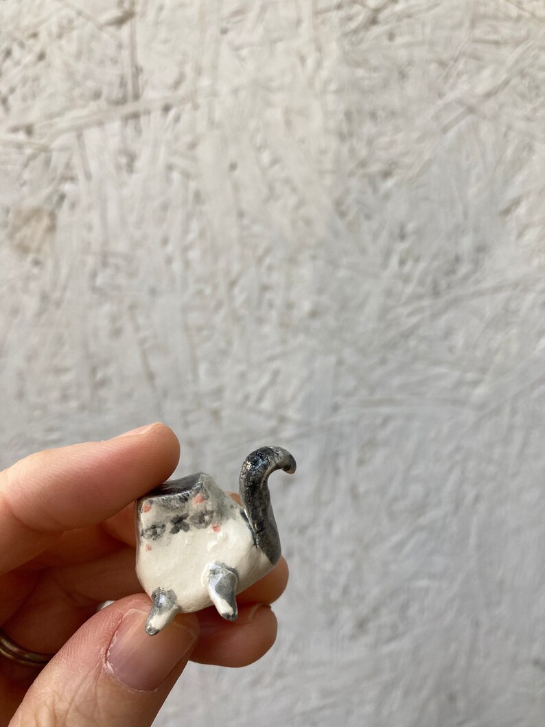May include: A small, handmade ceramic planter in the shape of a turtle. The planter is primarily white with black and gray accents, including the turtle's head and tail. The planter has three small feet and a small opening for planting.