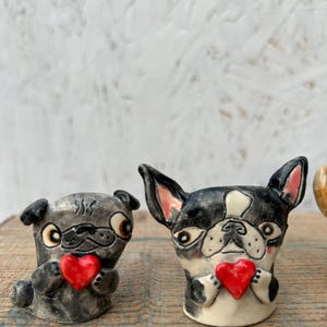 May include: Two ceramic dog figurines, a pug and a Boston terrier, each holding a red heart. The pug is gray with black accents, while the terrier is black and white. Both have expressive faces and are on a wooden surface.