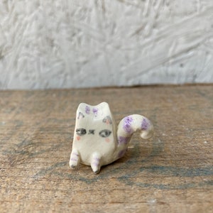 May include: A small, handmade ceramic cat figurine. The cat is white with purple markings on its head, legs, and tail. It has a whimsical design with closed eyes and a curled tail. The figurine is sitting on a wooden surface.