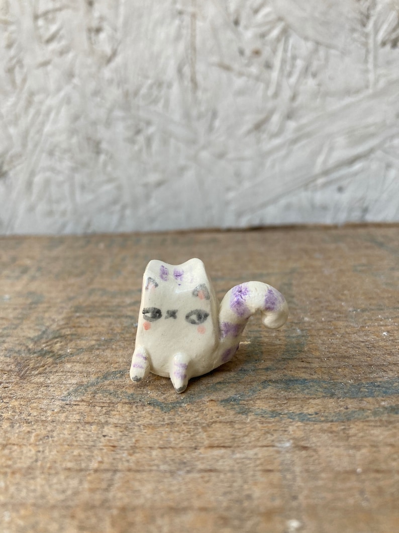 May include: A small, handmade ceramic cat figurine. The cat is white with purple markings on its head, legs, and tail. It has a whimsical design with closed eyes and a curled tail. The figurine is sitting on a wooden surface.