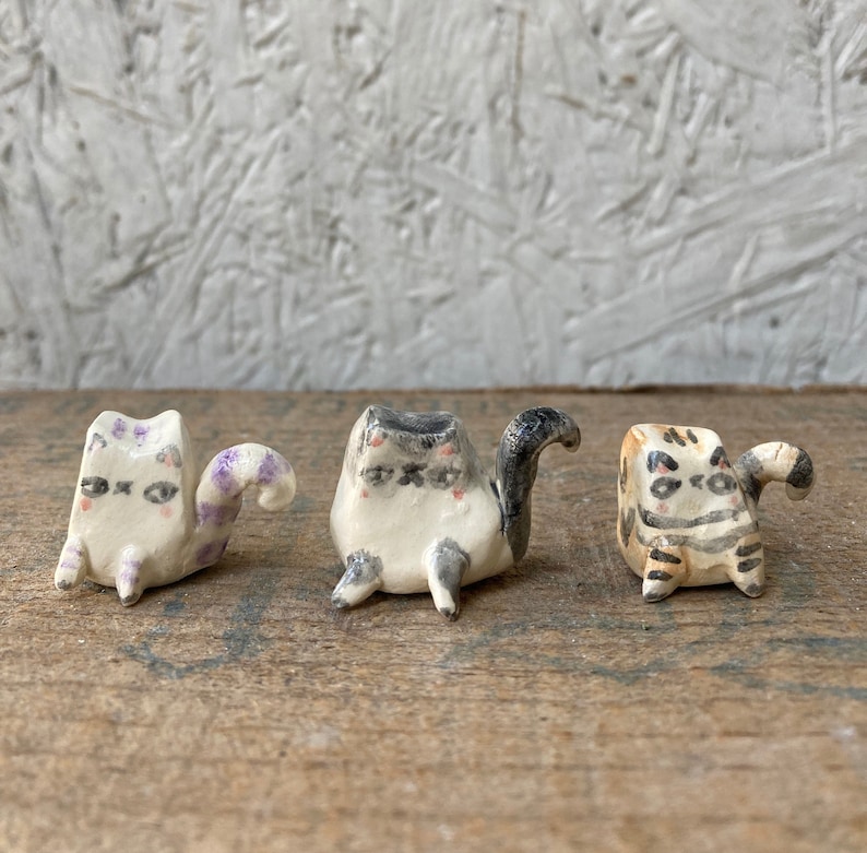 May include: Three handmade ceramic cat figurines. The cats are white with gray, purple, and tan markings. Each cat has a unique pose and expression, with small black eyes and pink cheeks. The figurines are on a wooden surface.