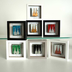 Könnte beinhalten: Ein Set aus neun gerahmten Glaskunstwerken mit einem stilisierten Kirchengebäude in verschiedenen Farben, darunter Gold, Blau, Rot, Grün und Schwarz. Jedes Stück ist in einem weißen, grauen oder schwarzen Rahmen gerahmt.