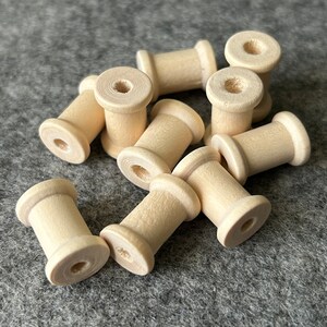 May include: A collection of small, unfinished wooden spools. Each spool has a cylindrical body with a central hole and flared ends. The spools are a light, natural wood colour and are scattered on a grey surface.