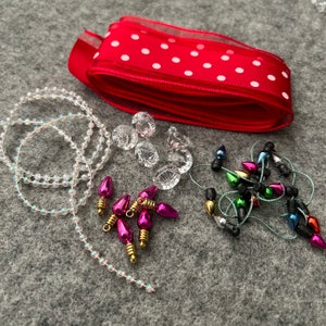May include: A collection of Christmas craft supplies on a gray surface. Includes a red ribbon with white polka dots, clear faceted gems, strings of clear beads, and miniature light bulbs in various colors.
