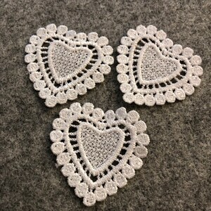 May include: Three white lace heart-shaped appliques. Each heart features a detailed, openwork centre and a scalloped edge. The appliques are arranged on a grey surface, showcasing their delicate design and texture. Suitable for crafting or embellishing projects.