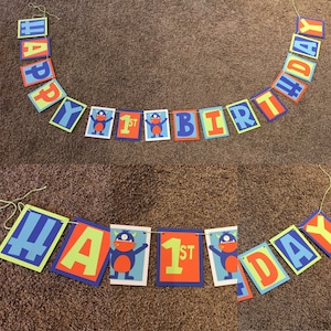 May include: A colorful banner with the words "HAPPY BIRTHDAY" strung across a string. The banner includes individual rectangular flags with letters in blue, green, orange, and red. Some flags feature a cartoon monster and the number "1st".