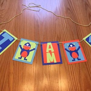May include: A colorful banner with the text "I AM 1" strung on a yellow cord. The banner features blue, green, and orange rectangles with letters and monster illustrations. The number 1 is in blue.