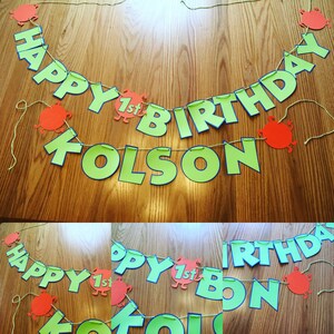 May include: A handmade birthday banner with the words "HAPPY BIRTHDAY KOLSON" in large, green letters with blue outlines. The banner also includes the text "1st" and orange decorative cutouts. The banner is strung on a thin, yellow string.