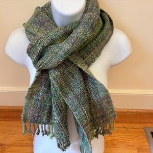 May include: A hand-woven scarf featuring a variegated blend of green, olive, and subtle purple yarns. The textile has a textured, loose-weave appearance with visible cross-hatching and short, frayed fringe detailing along the bottom edges.