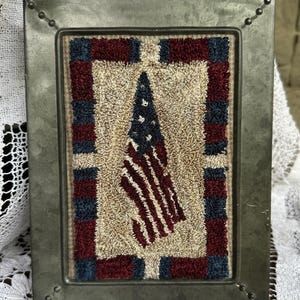 Peut inclure: Un cadre métallique rectangulaire à la finition gris foncé contenant une œuvre d'art en tapis crocheté. Le tapis présente un motif central de drapeau américain en rouge, blanc et bleu, entouré d'une bordure de carrés rouges et bleus alternés.