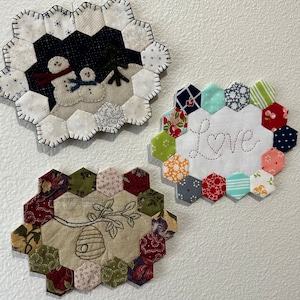 May include: Three hexagon-shaped quilted wall hangings with different designs. The first features a snowman family with a dark blue background. The second has a white background with the word "Love" embroidered in a cursive font with a heart. The third has a beehive and a branch with leaves embroidered on a beige background.