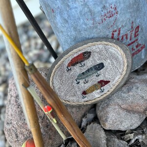 May include: A close-up of a wooden circle with three colorful fishing lures embroidered on it. The circle is surrounded by a rope border. A fishing rod and a metal bucket are in the background.
