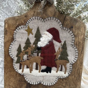 May include: A rustic wooden cutting board with a decorative felt applique featuring Santa Claus, reindeer, and Christmas trees. The applique is framed with a scalloped edge and a black and white checkered bow at the top. A gold star is also included in the design.
