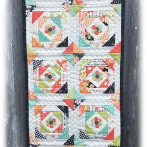 May include: A colorful patchwork table runner with a black border. The runner features a repeating pattern of squares with a floral design in the center. The colors used are white, black, orange, green, and pink.