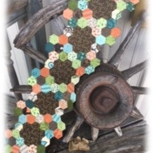 May include: A long table runner made of fabric hexagons in shades of brown, green, orange, and blue. The hexagons are sewn together to create a continuous pattern.