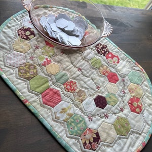 May include: A white oval quilted table runner with a green border and a hexagonal pattern. The runner is decorated with various floral and geometric prints in shades of red, green, brown, and cream. A glass bowl filled with white paper hexagons sits on top of the runner.