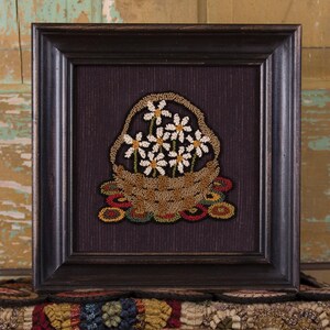 May include: Framed artwork featuring a basket of white daisies with yellow centers. The basket is brown, and the background is a dark purple. The frame is dark brown. The artwork is a needlepoint or rug hooking design.