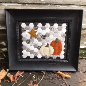 May include: A framed felt wall hanging with a patchwork of gray and white hexagons, a gold star, and two felt pumpkins, one orange and one white.