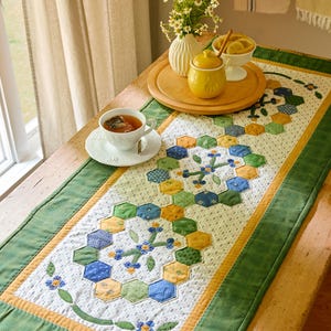 May include: A handmade table runner with a patchwork design featuring hexagonal shapes in blue, green, and yellow. The runner is bordered with green fabric and has floral accents. A cup of tea and a small vase are on the table.