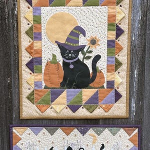 May include: A rectangular fabric wall hanging featuring a black cat wearing a purple witch hat, sitting between two orange pumpkins under a yellow moon. A smaller horizontal fabric piece below shows a black cat, pumpkins, and a white fence.