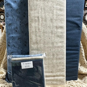 May include: Three bolts of fabric standing upright: one blue with a dark floral pattern, one cream with a geometric lattice design, and one dark blue with a fine speckled texture. A plastic-wrapped bundle of the same fabrics sits in front.