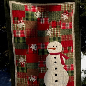 May include: A patchwork quilt with a snowman design. The quilt is made of red, green, and brown plaid fabric. The snowman is white with black eyes and buttons, a carrot nose, and a red scarf.