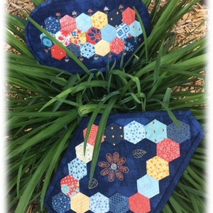 May include: Two blue quilted placemats with a hexagon patchwork design. Each placemat features a border of colorful fabric hexagons. One placemat has a floral design in the center. The placemats are set against a background of green grass.