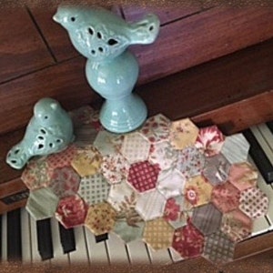 May include: A teal blue ceramic bird figurine sits on a hexagonal patchwork quilt. The quilt is made of various fabrics in shades of red, white, yellow, and green. The quilt is on top of a piano keyboard.