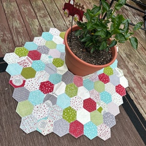May include: A multi-colored patchwork quilt in the shape of a Christmas tree with a red truck ornament on top. The quilt is made of various fabrics with different patterns, including red, green, white, and blue. The quilt is sitting on a wooden deck with a potted plant in the background.